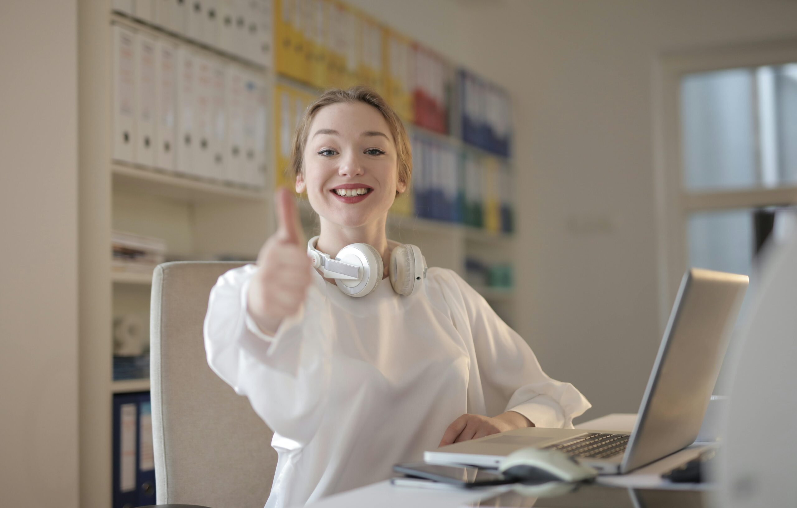 Smiling woman giving thumbs up while working with laptop and headphones in an office.